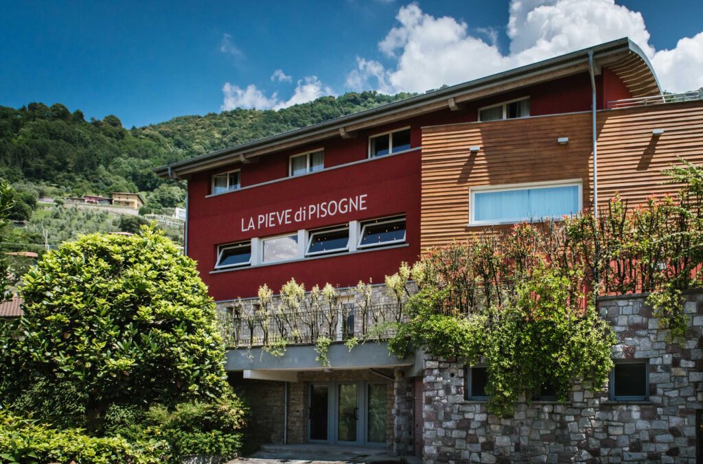 Edificio moderno con facciata in legno e muratura, situato in un ambiente collinare verde.