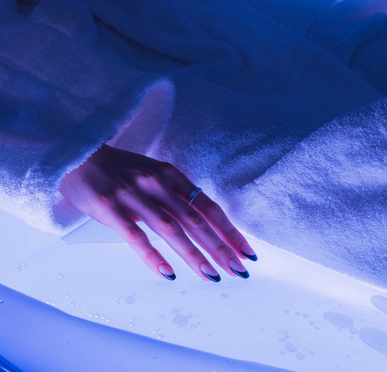 Eine Hand mit dunklem Nageldesign ruht auf einer nassen, beleuchteten Oberfläche.