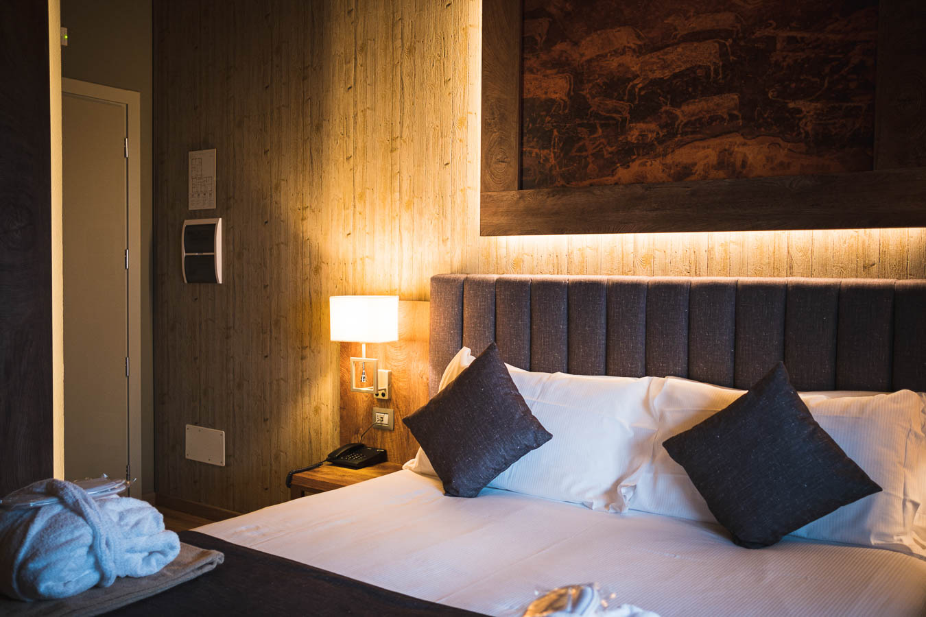 Camera d'albergo accogliente con letto matrimoniale, cuscini scuri e lampada da comodino illuminata.