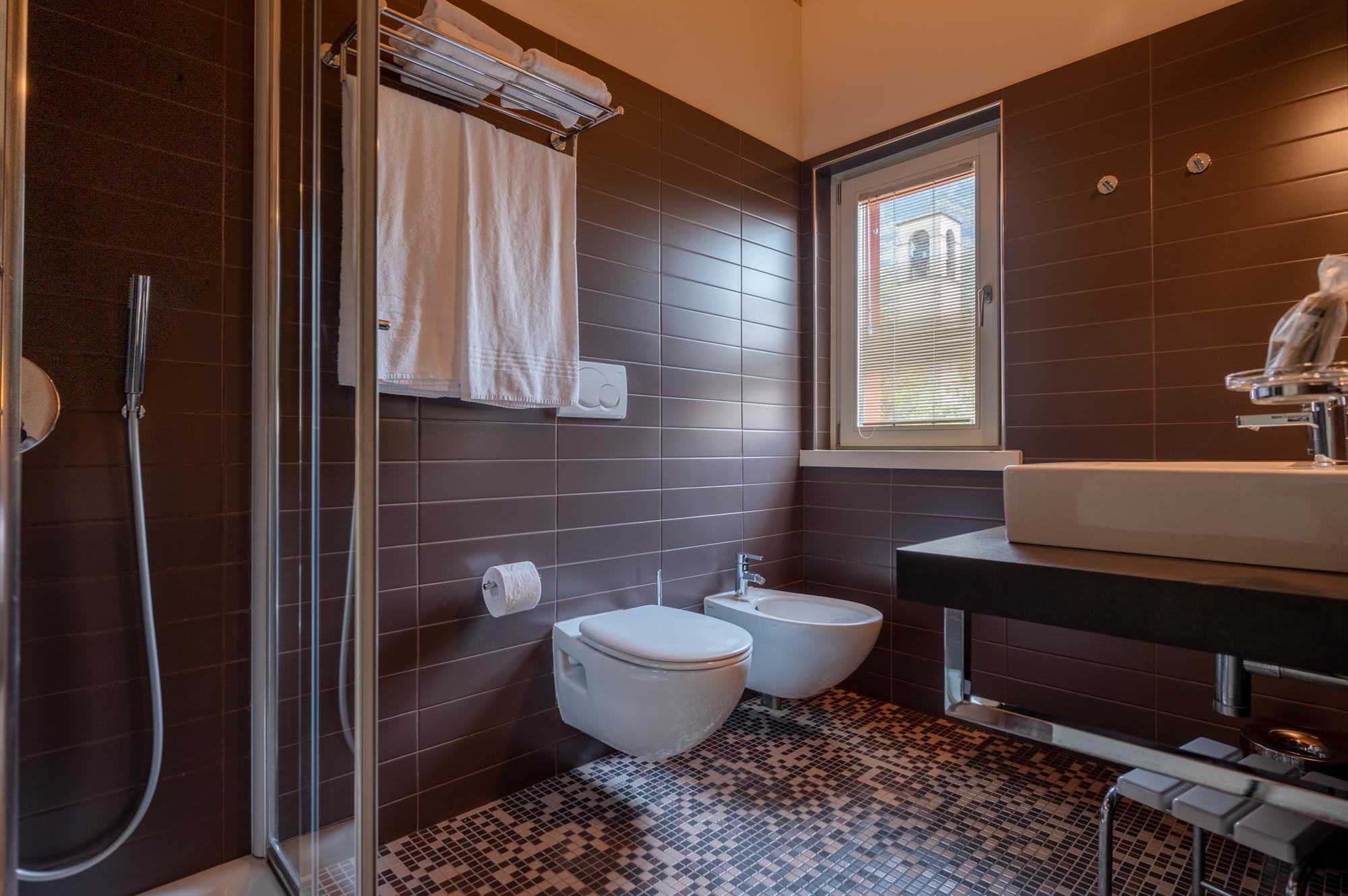 Bagno moderno con piastrelle marroni, doccia, lavabo, bidet e finestra.