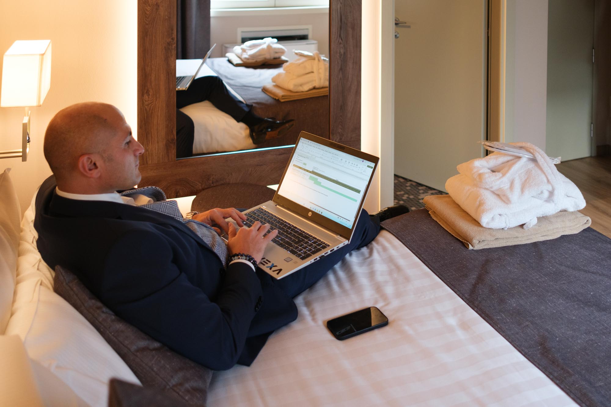 Uomo in abito lavora al computer portatile sdraiato su un letto in hotel.