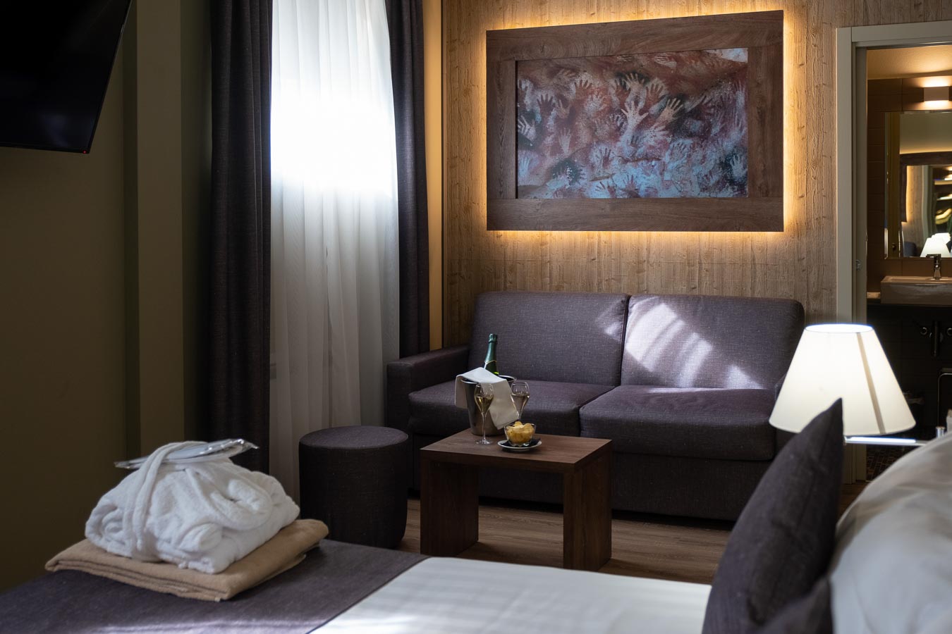 A cozy hotel room with a sofa, artwork, and a small table holding a bottle and glasses.