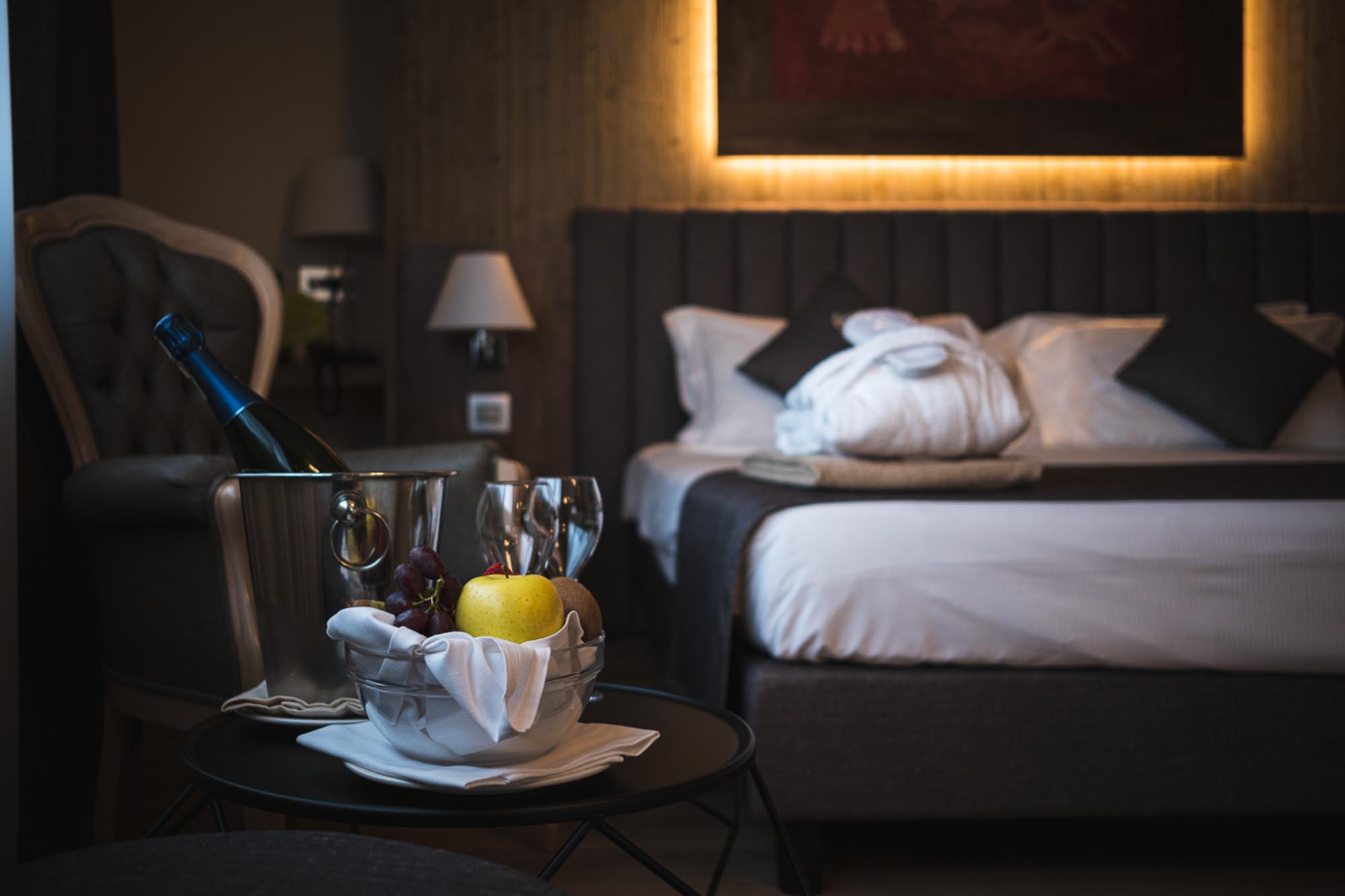 Camera d'albergo elegante con letto matrimoniale, frutta, spumante e bicchieri su un tavolino.