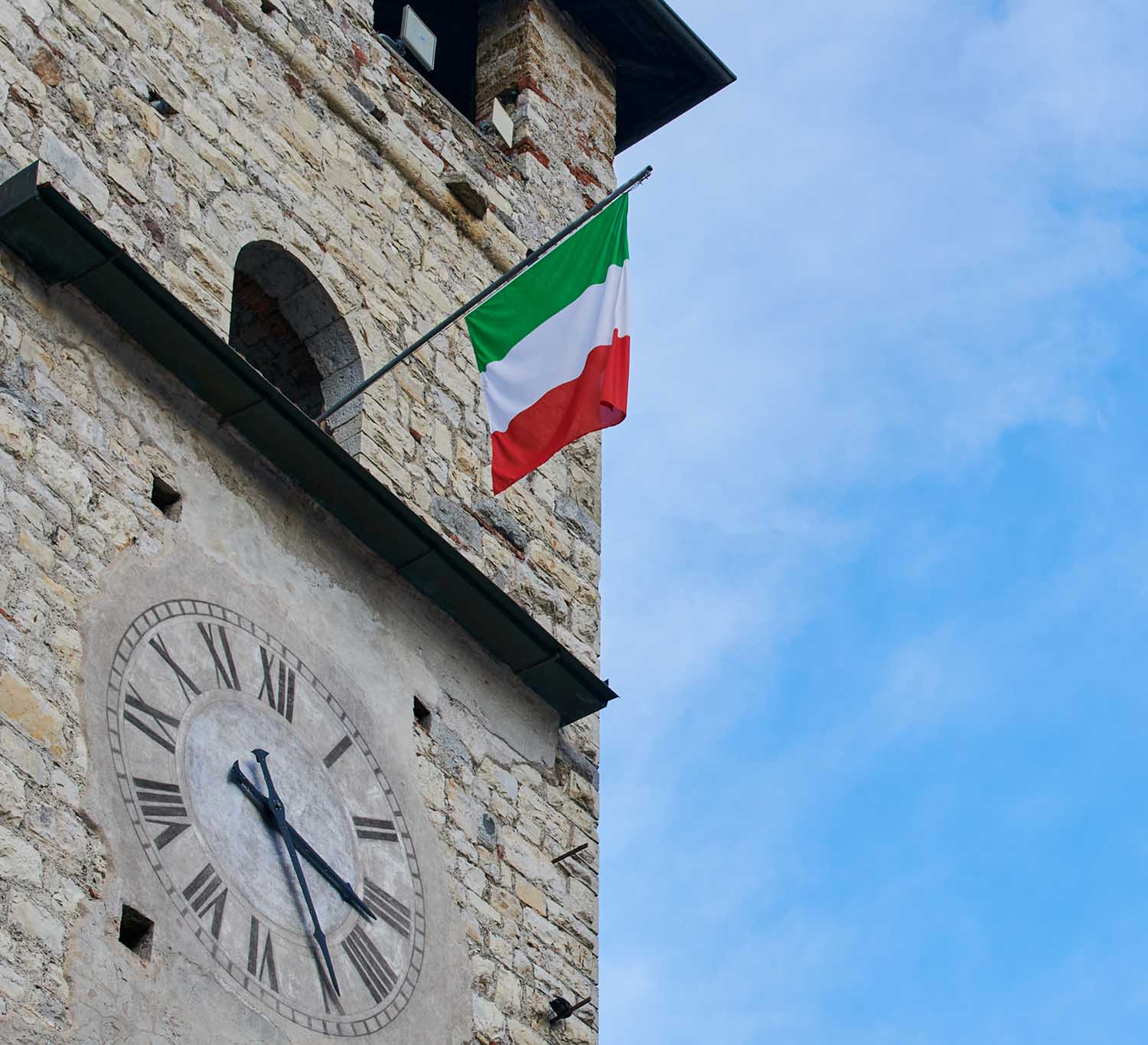 La facciata di un edificio storico con un orologio e una bandiera italiana su sfondo di cielo azzurro.