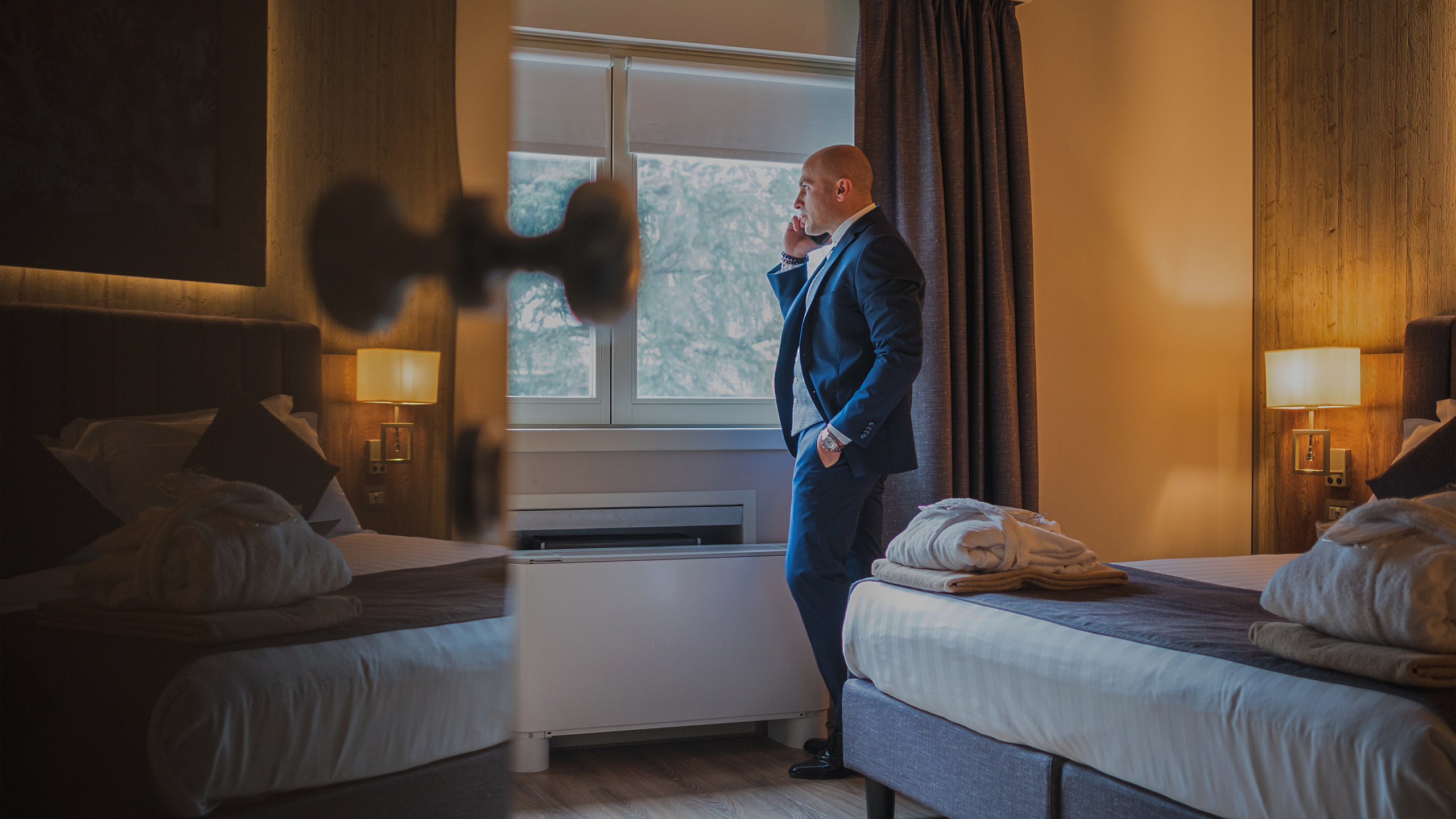 Uomo in abito che parla al telefono vicino alla finestra di una stanza d'albergo.