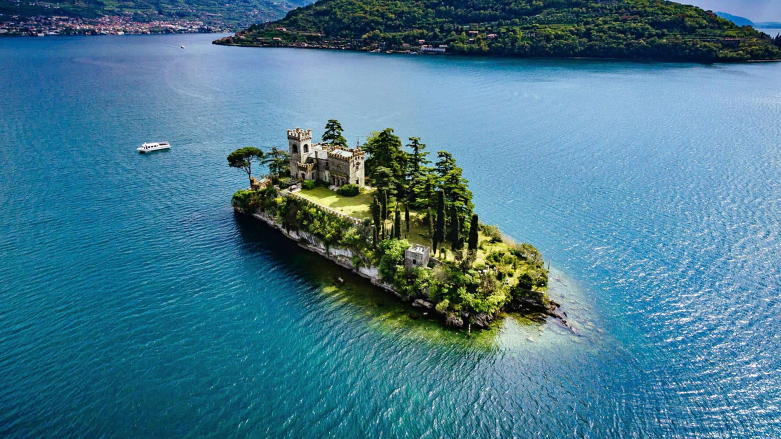 Veduta aerea dell'Isola di Loreto sul Lago d'Iseo con un castello circondato da alberi e acqua blu.